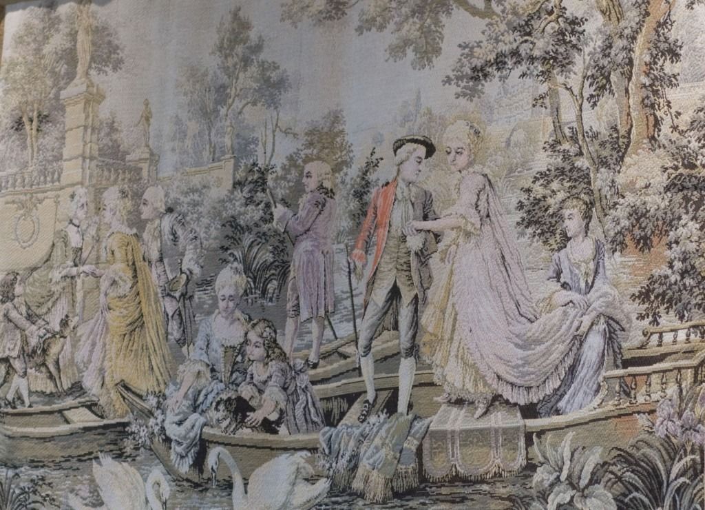 People in 18th-century dress gathered in a garden, with a seated woman at right and figures near a fountain.