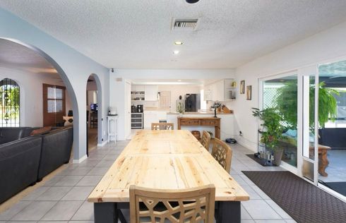 Bright open kitchen and dining area with a long wooden table, white cabinets, and sliding glass doors.