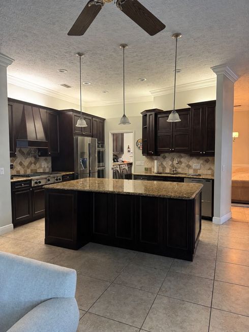 Spacious kitchen with dark wood cabinets, granite island, tile floor, and ceiling fans under warm lighting