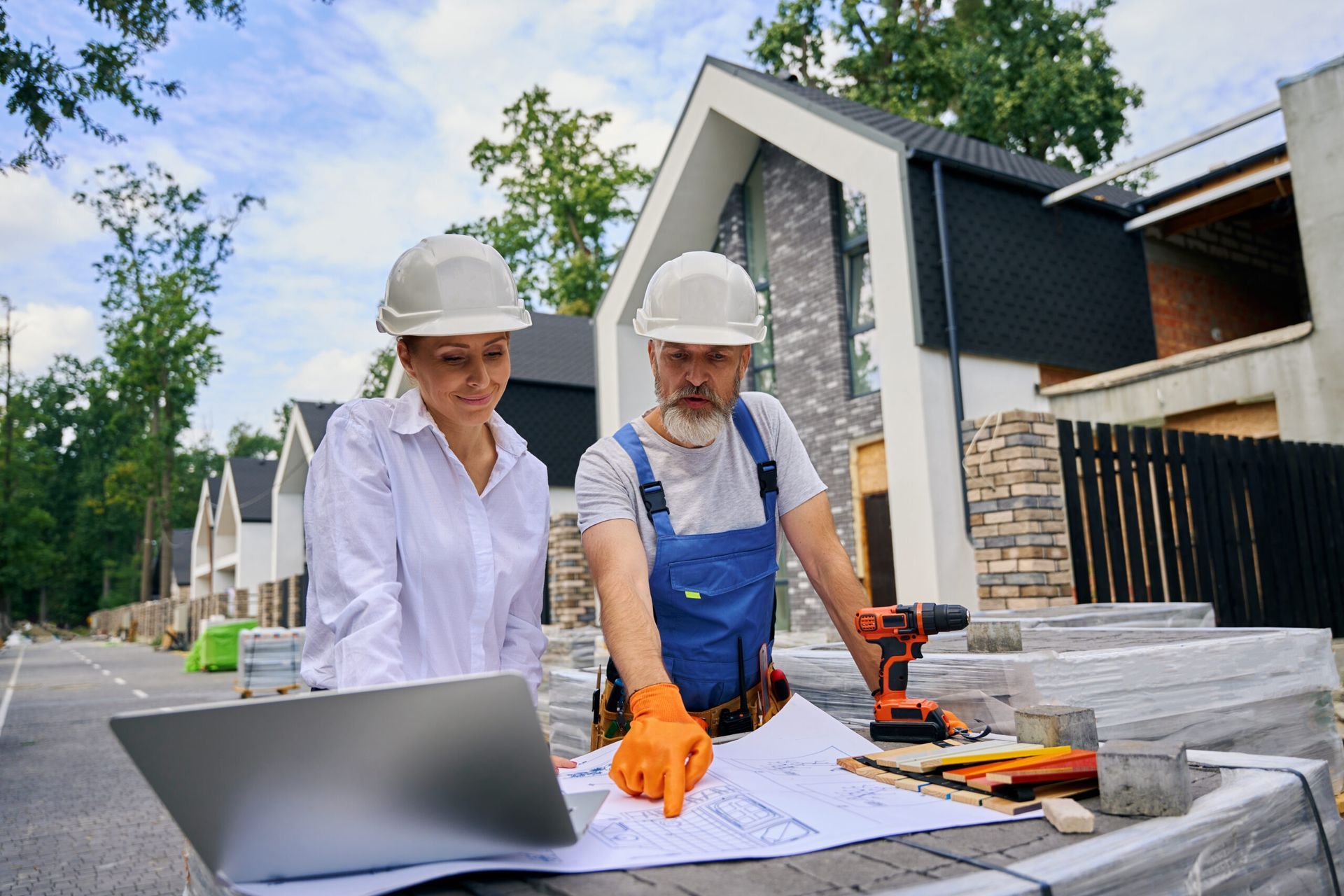 Two workers in hard hats review blueprints beside a laptop at an outdoor construction site.