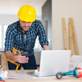 Construction worker in yellow hard hat measuring plans beside a laptop at a worksite