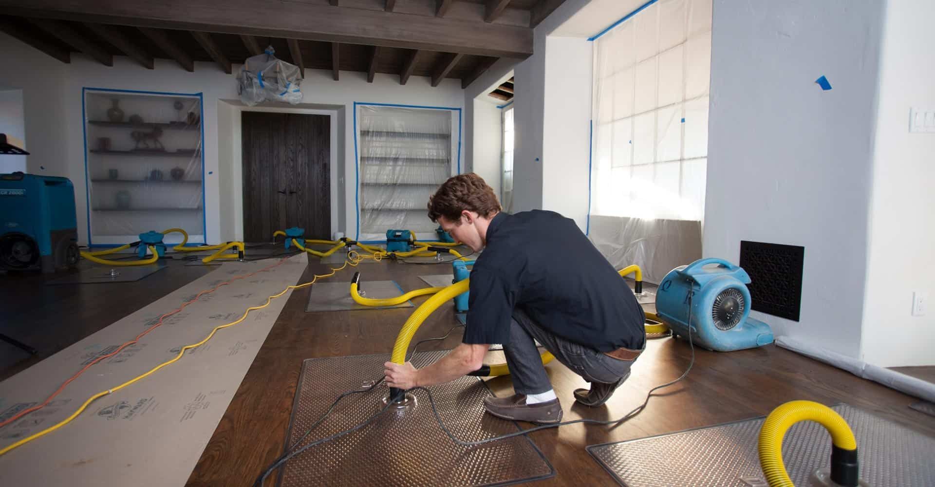 Person kneeling on a flooded floor, working with yellow hoses in a room with a large window.