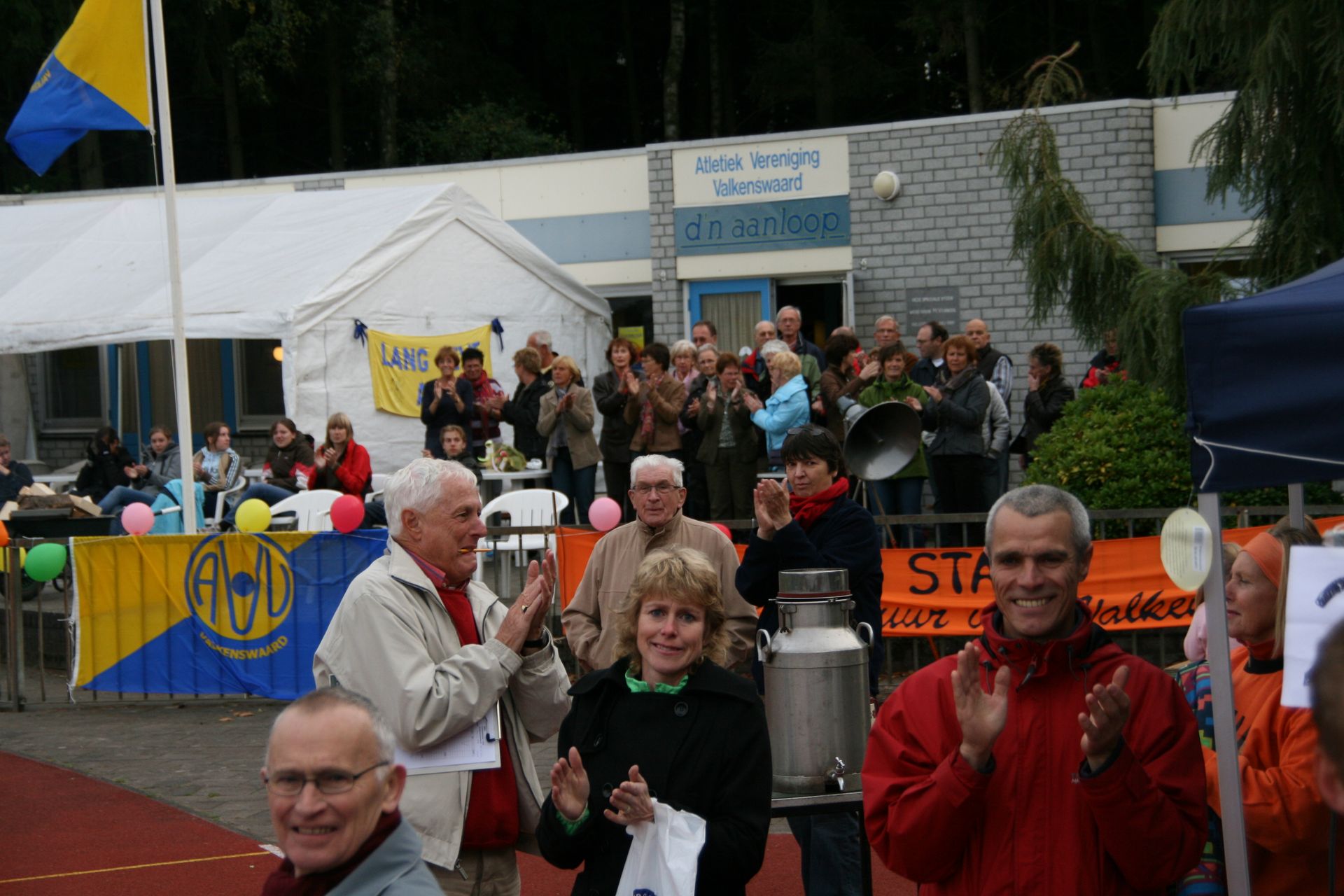 Mensen applaudisseren tijdens een openluchtevenement, met een gebouw en een tent op de achtergrond. 