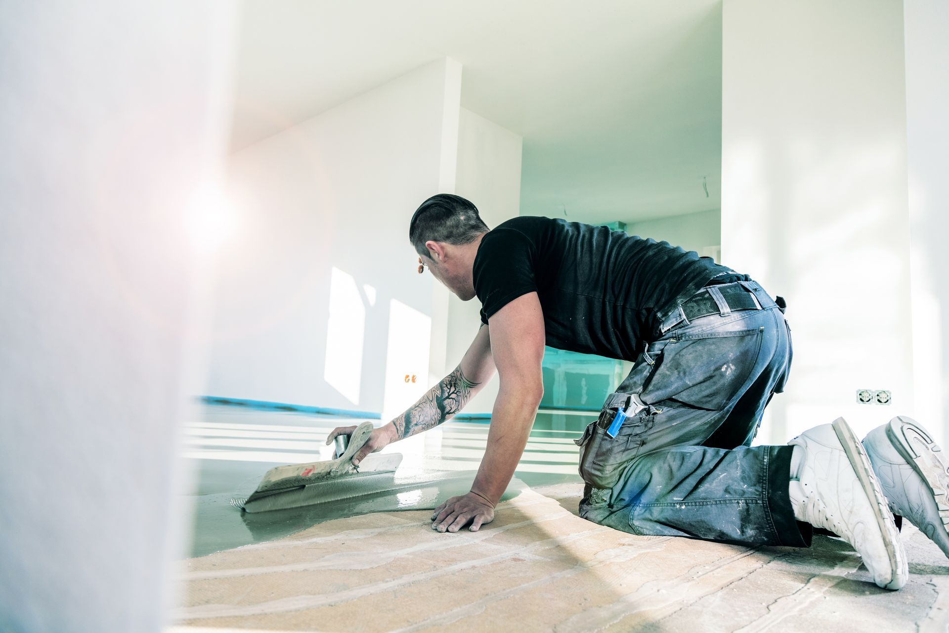 A person kneels on a light-colored floor, smoothing wet cement with a trowel in a brightly lit, unfinished interior space.