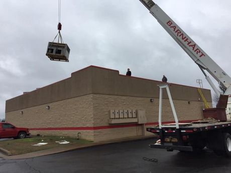 A crane lifts an HVAC unit onto a low commercial building roof. Two workers stand on the roof.