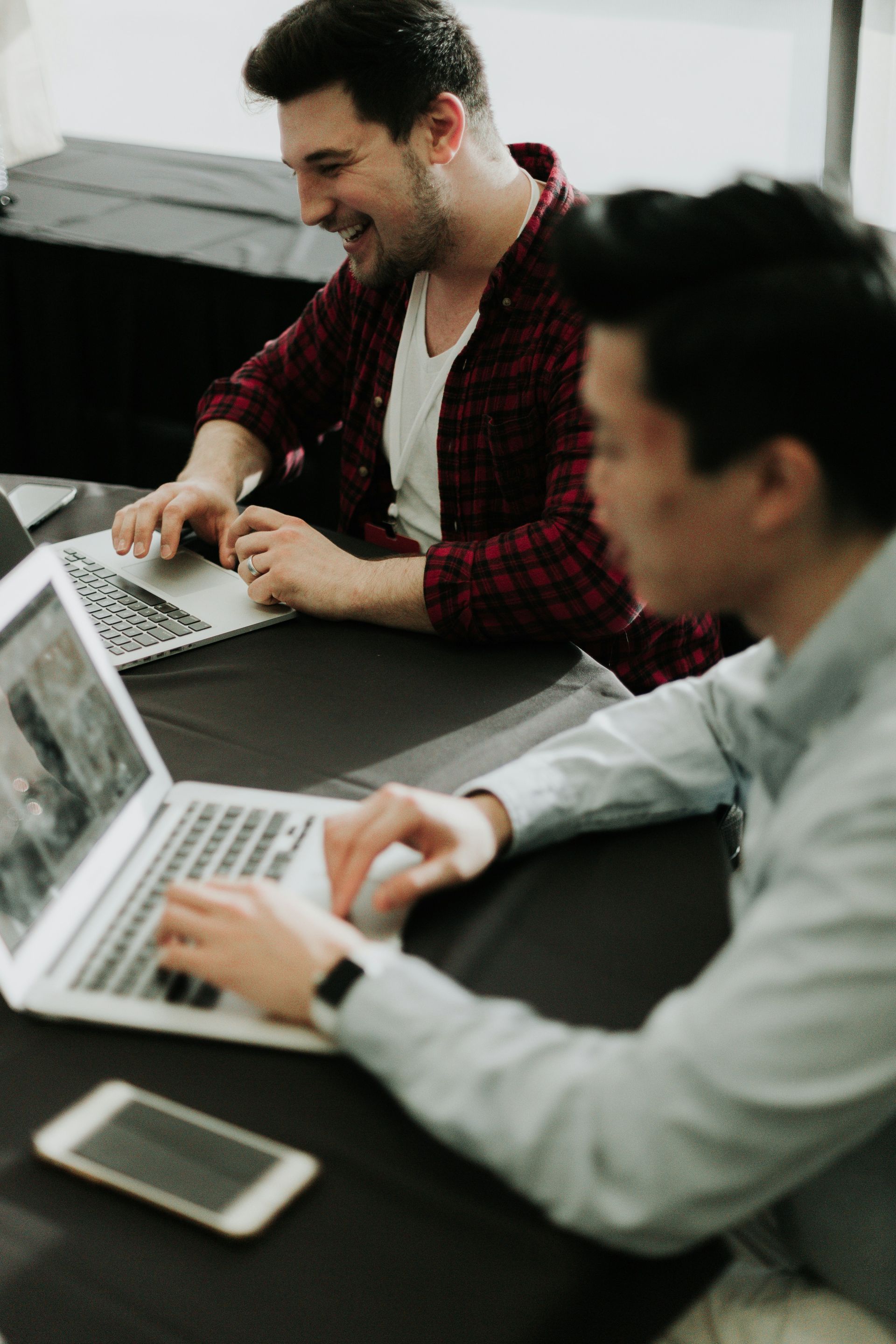 Two coworkers using laptops at a table, smiling in a bright office.