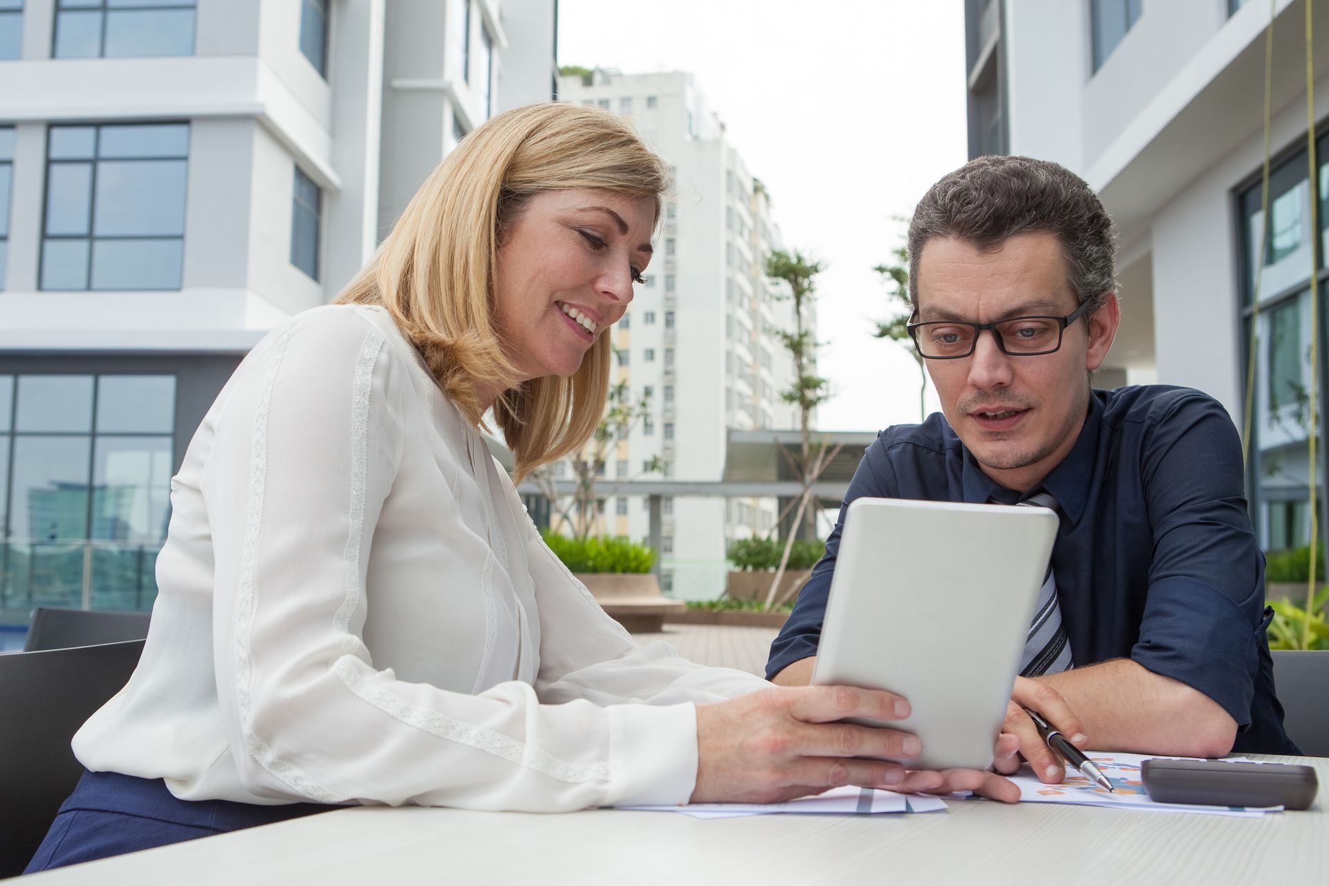 Two people reviewing a tablet together at an outdoor table between office buildings