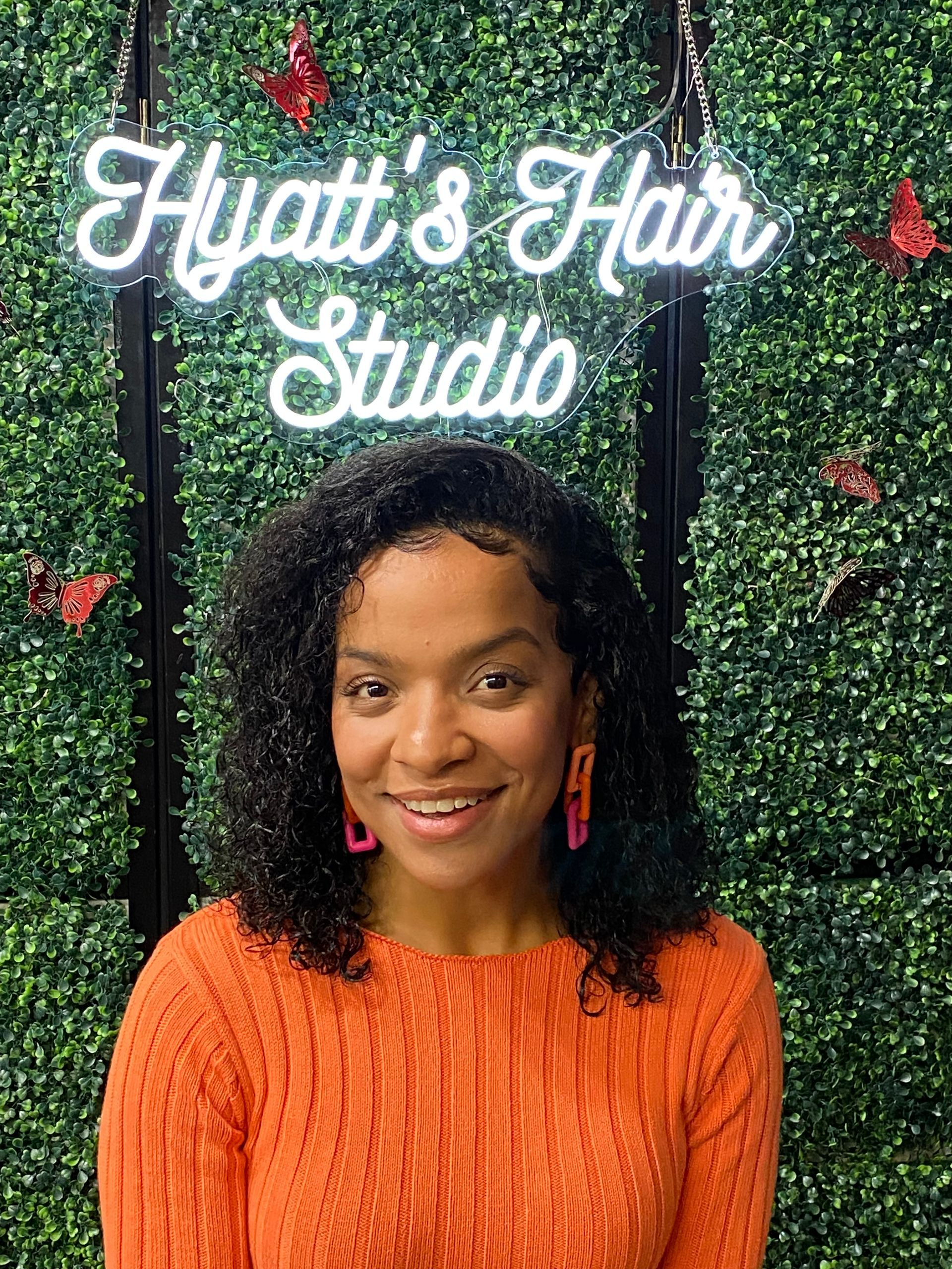 A woman is smiling in front of a hyatt 's hair studio sign.