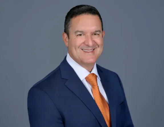 Man in navy suit and orange tie smiling against a gray studio background
