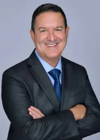 Smiling man in a dark suit and blue tie, arms crossed against a light gray background