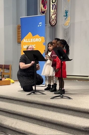 A group of young girls are singing on a stage in front of a sign that says allegro music.