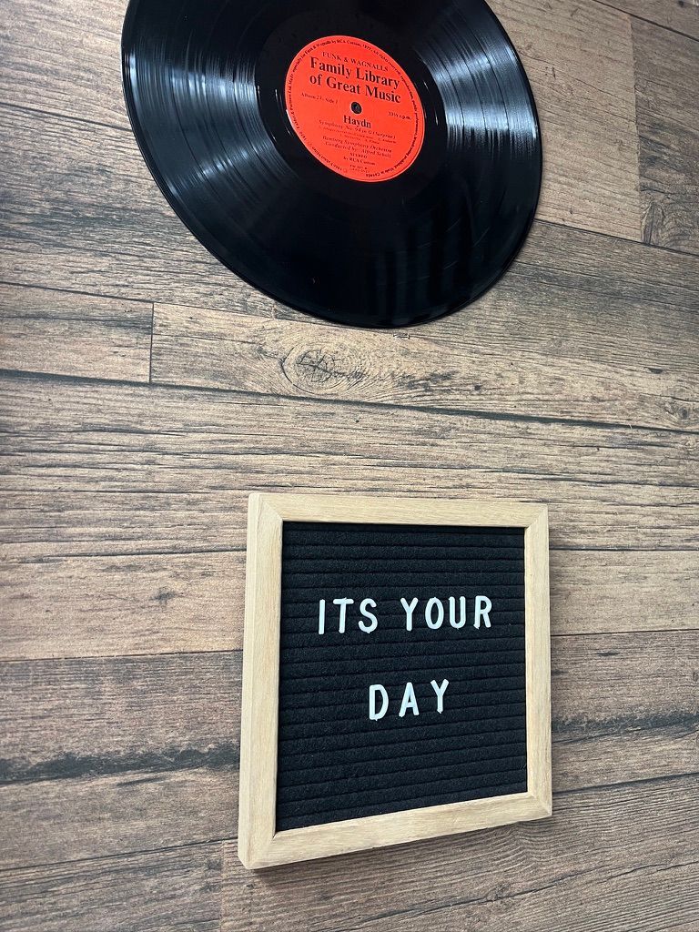 A record is hanging on a wooden wall next to a letter board that says `` it 's your day ''.