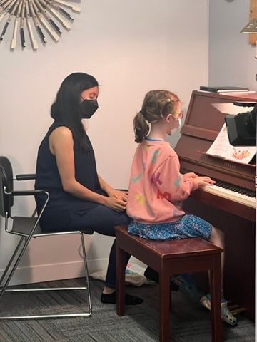 A woman wearing a mask is teaching a little girl how to play the piano.