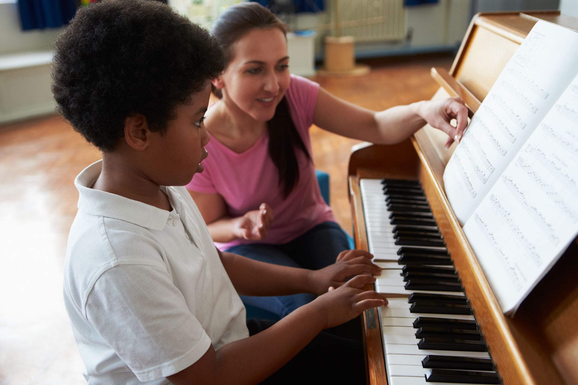 A young Black boy learning to play the piano with a smiling female instructor in a classroom.