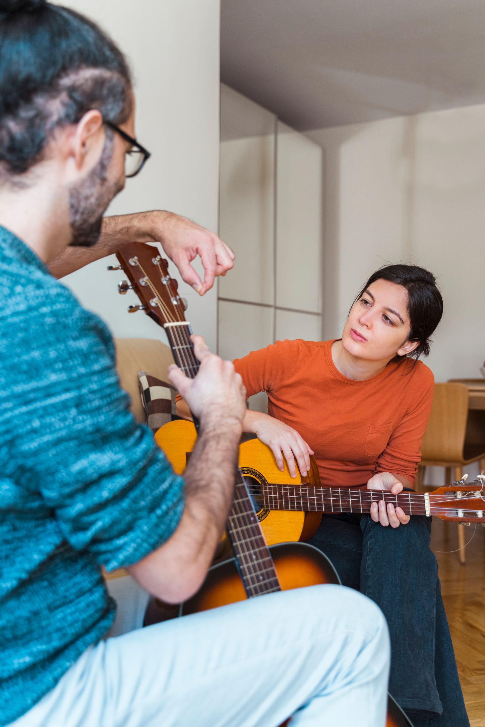 Two people playing acoustic guitars in a bright living room, one seated and smiling.