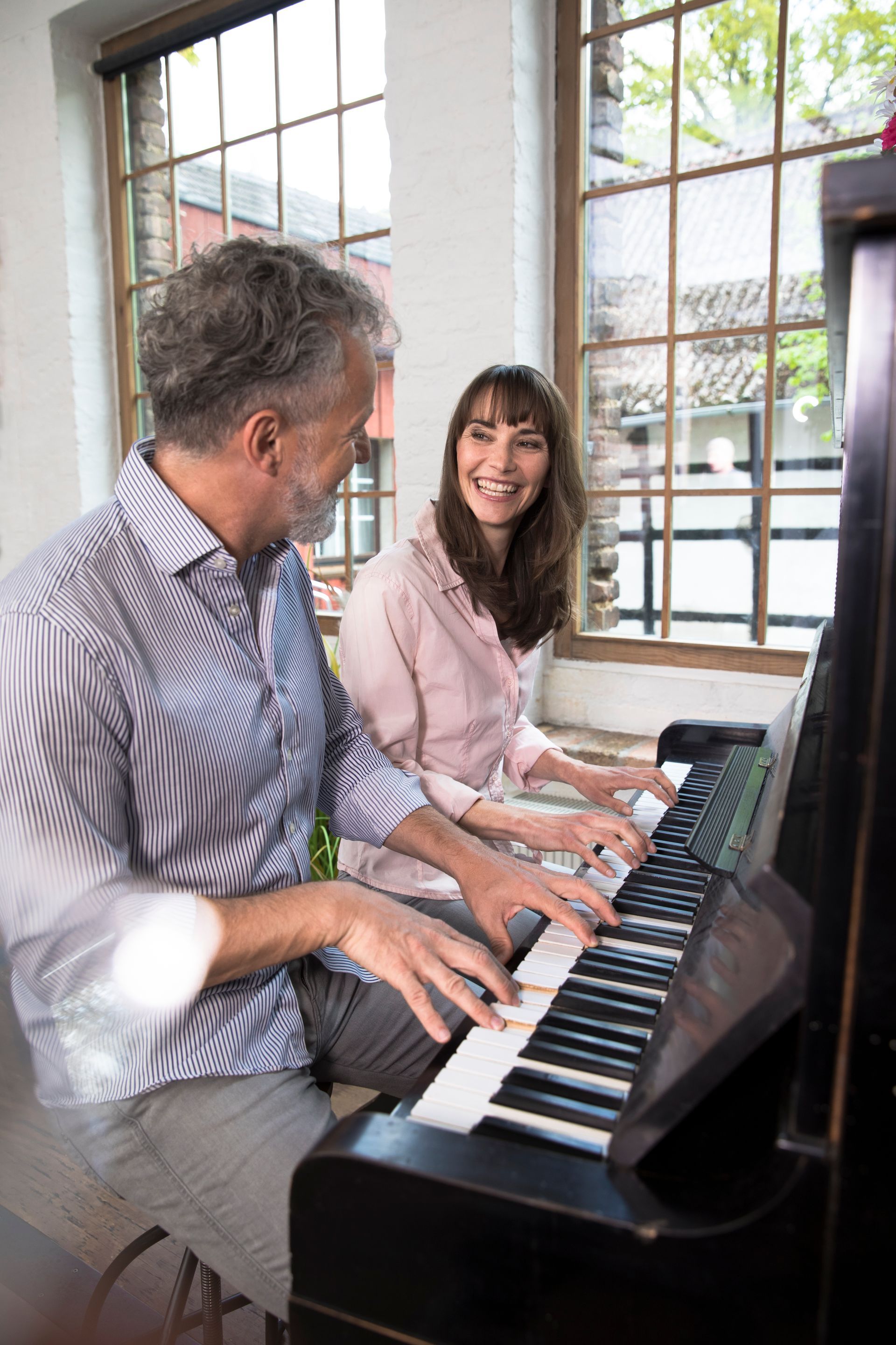 Two people smiling while playing a black piano in a bright room with large windows