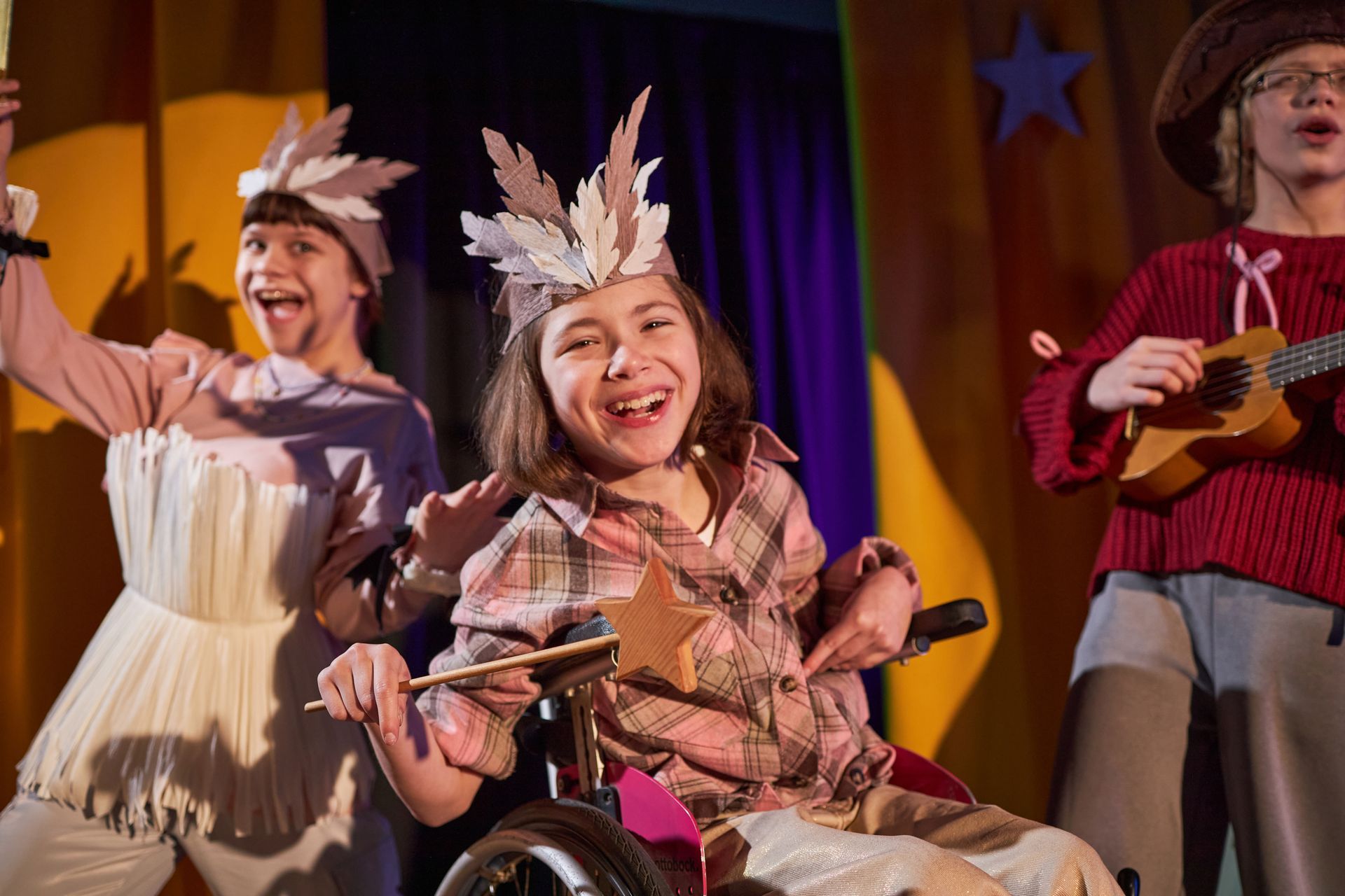 Children in festive costumes performing on stage, smiling under warm purple and orange lights.