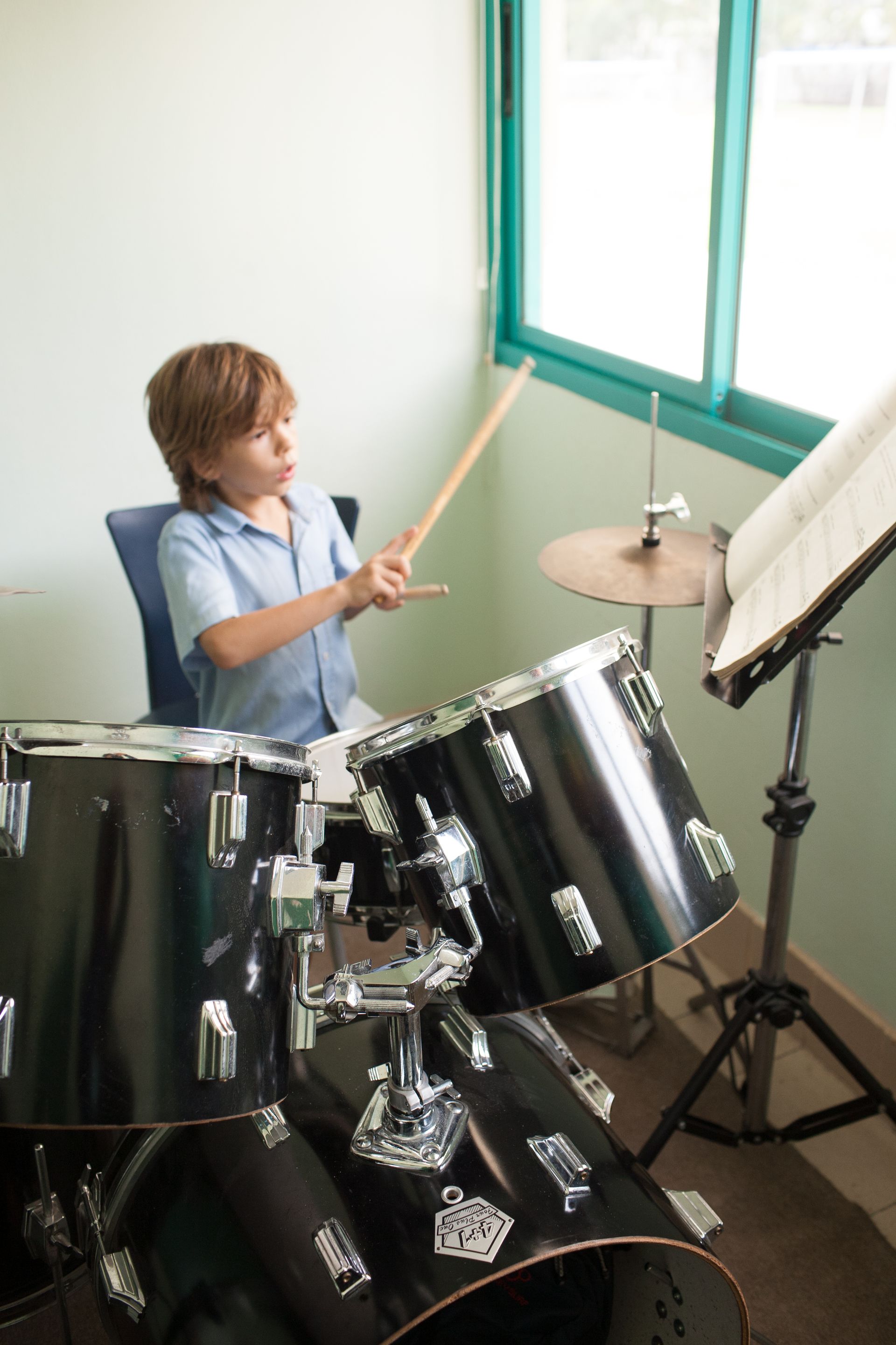 Young person playing a black drum set, hitting drums with sticks. Sheet music on stand by a window.