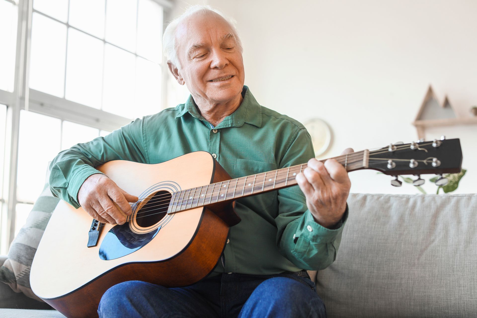 An elderly man is sitting on a couch playing an acoustic guitar.
