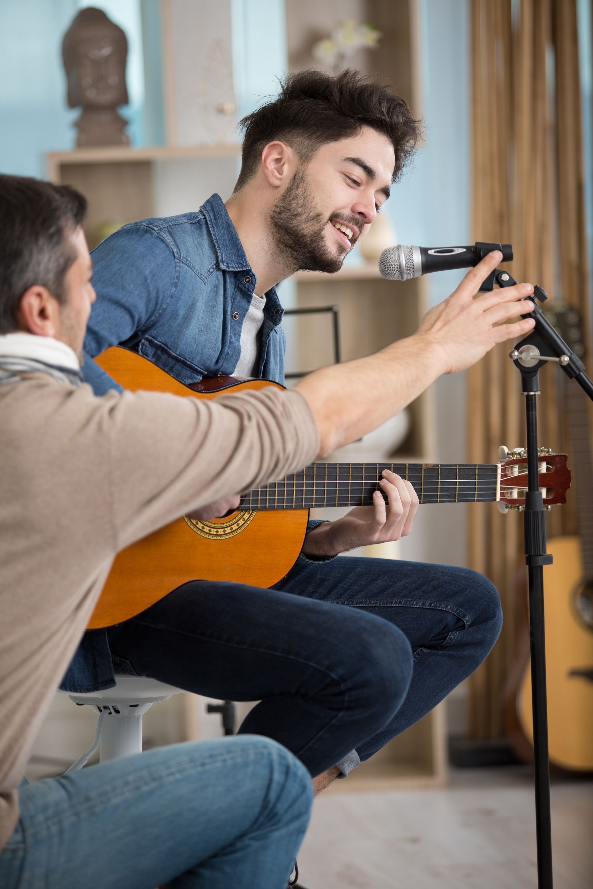 A man is teaching another man how to play a guitar and sing into a microphone.