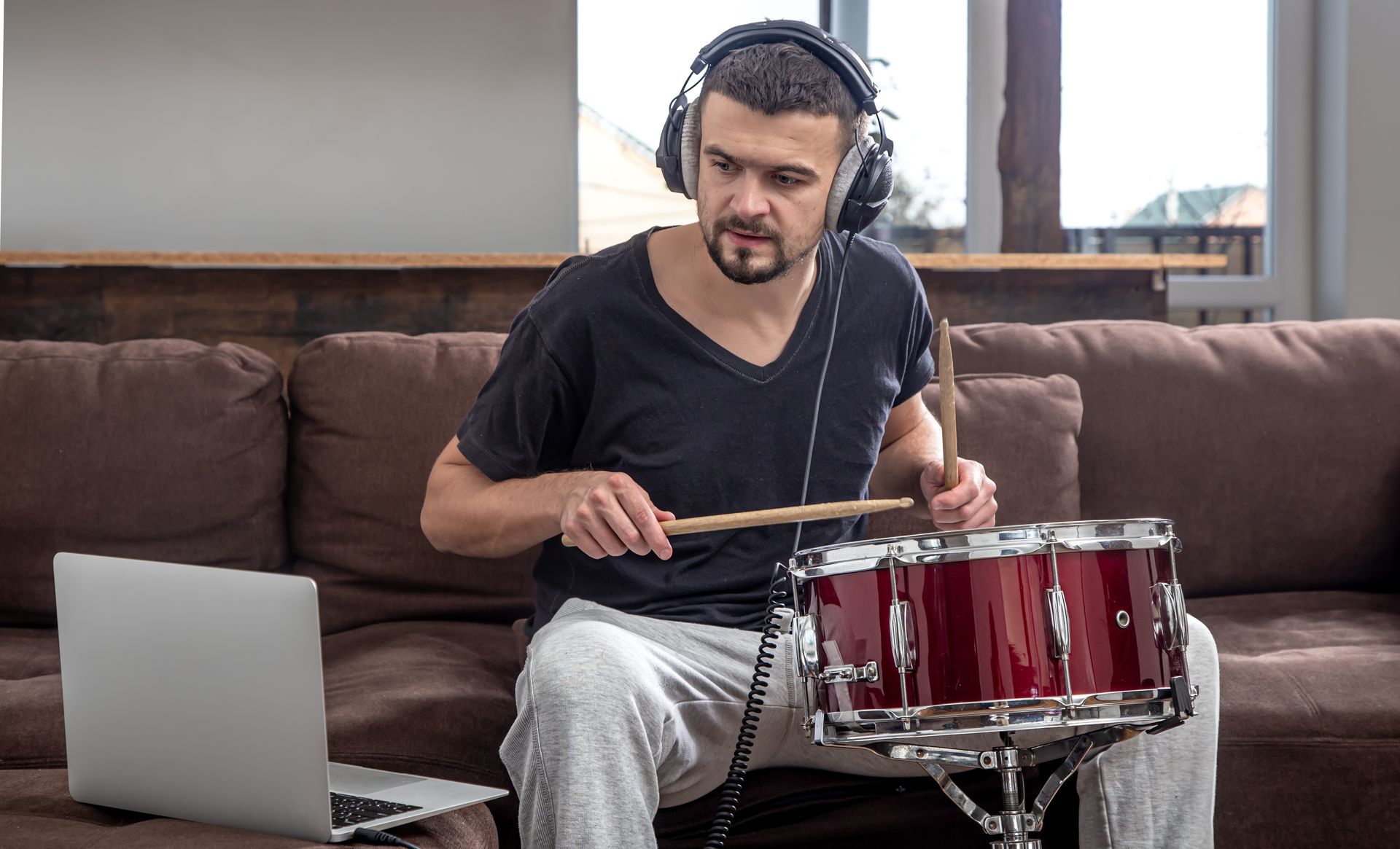 Man playing a red drum, wearing headphones, seated on a couch with a laptop.