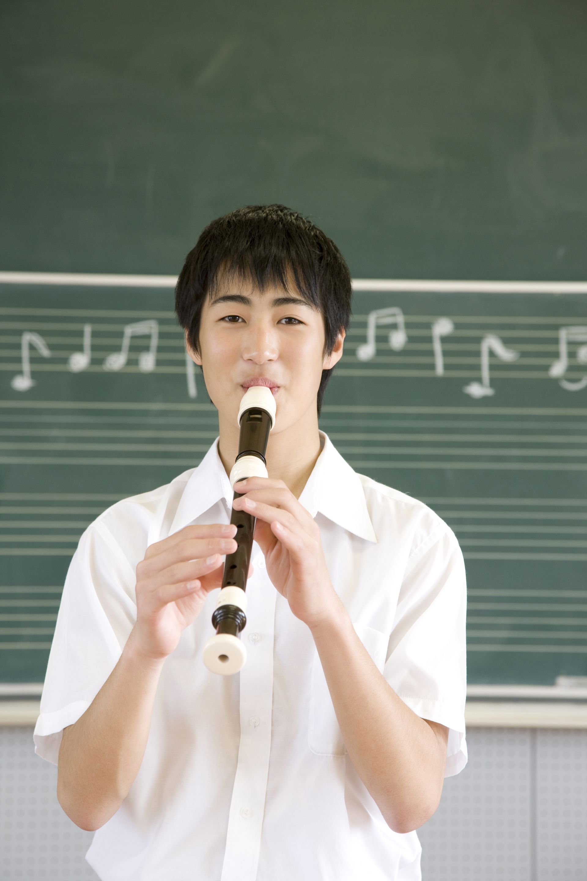 A young man is playing a recorder in front of a blackboard with music notes on it.