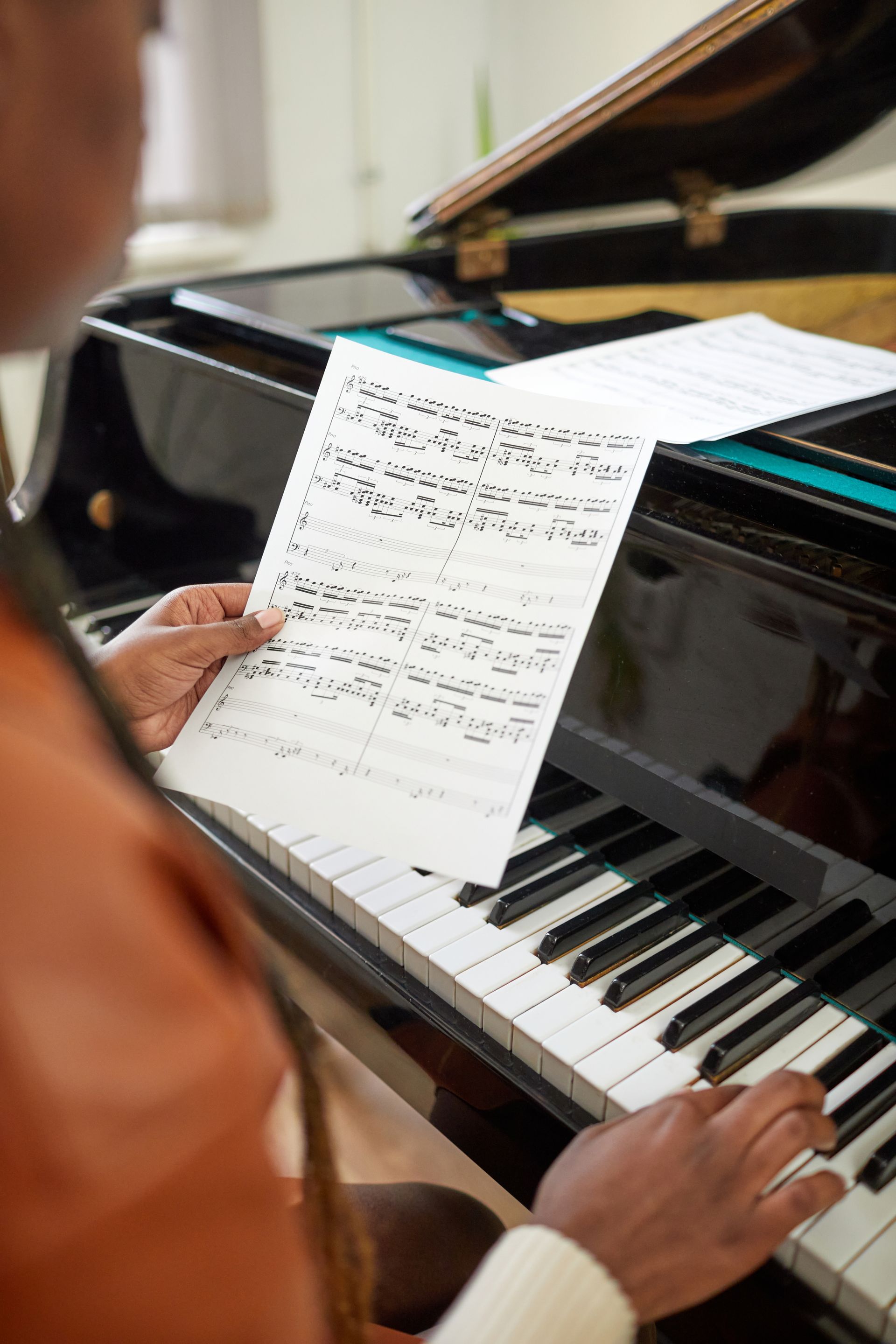 Person playing piano, holding sheet music. Black piano with keys, sheet music visible.