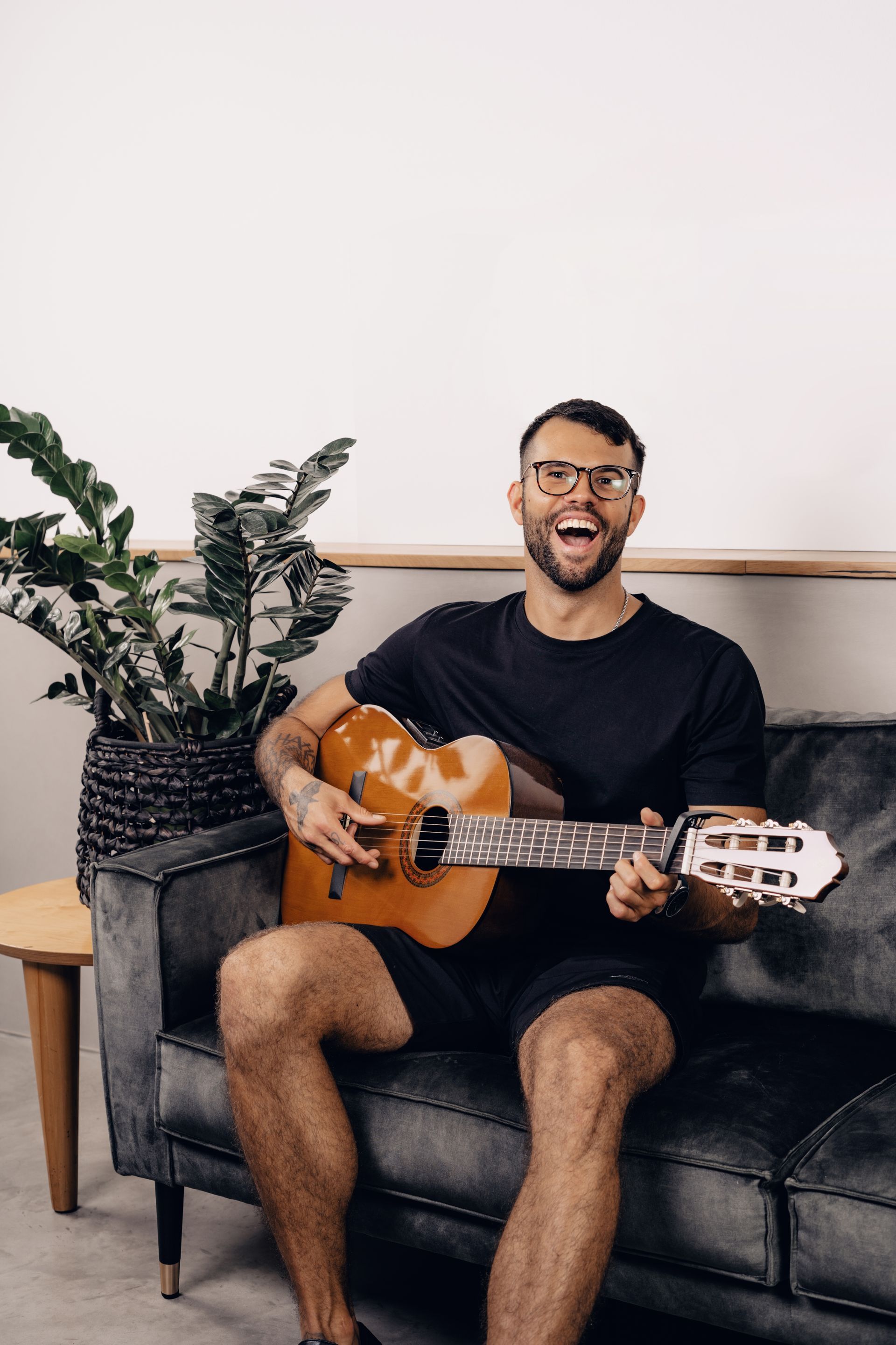Man playing acoustic guitar, smiling widely, sitting on a gray couch near a potted plant.