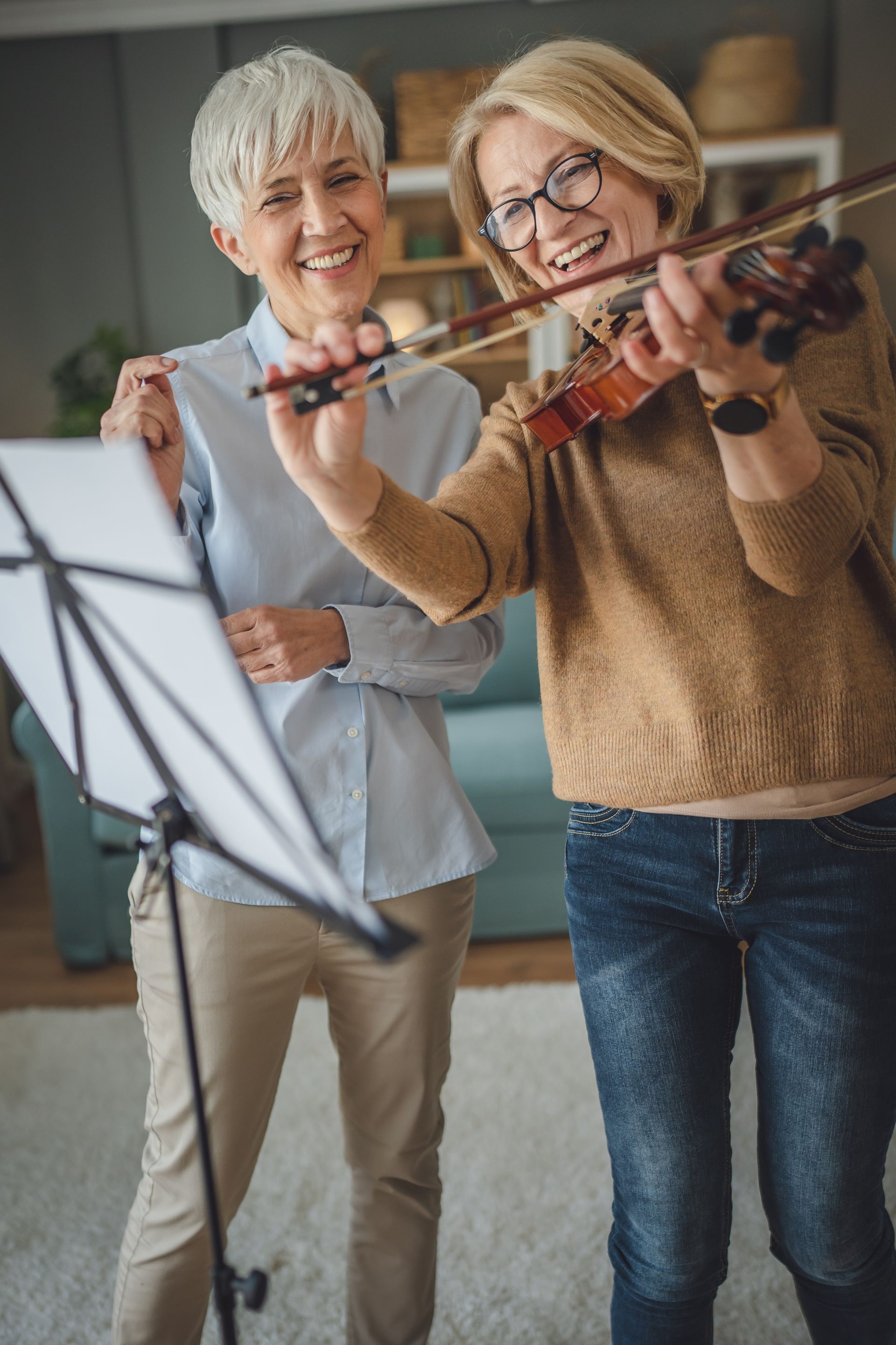 Two women in a room; one plays a violin, the other points at sheet music, smiling.