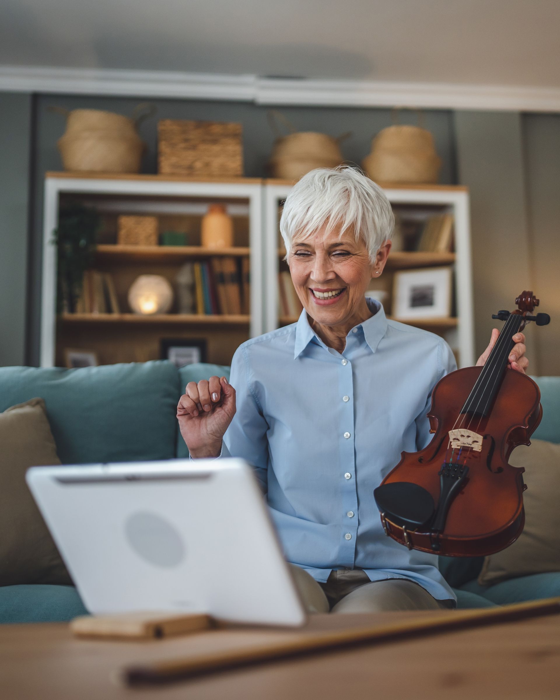 An elderly woman is holding a violin while sitting on a couch in front of a laptop.