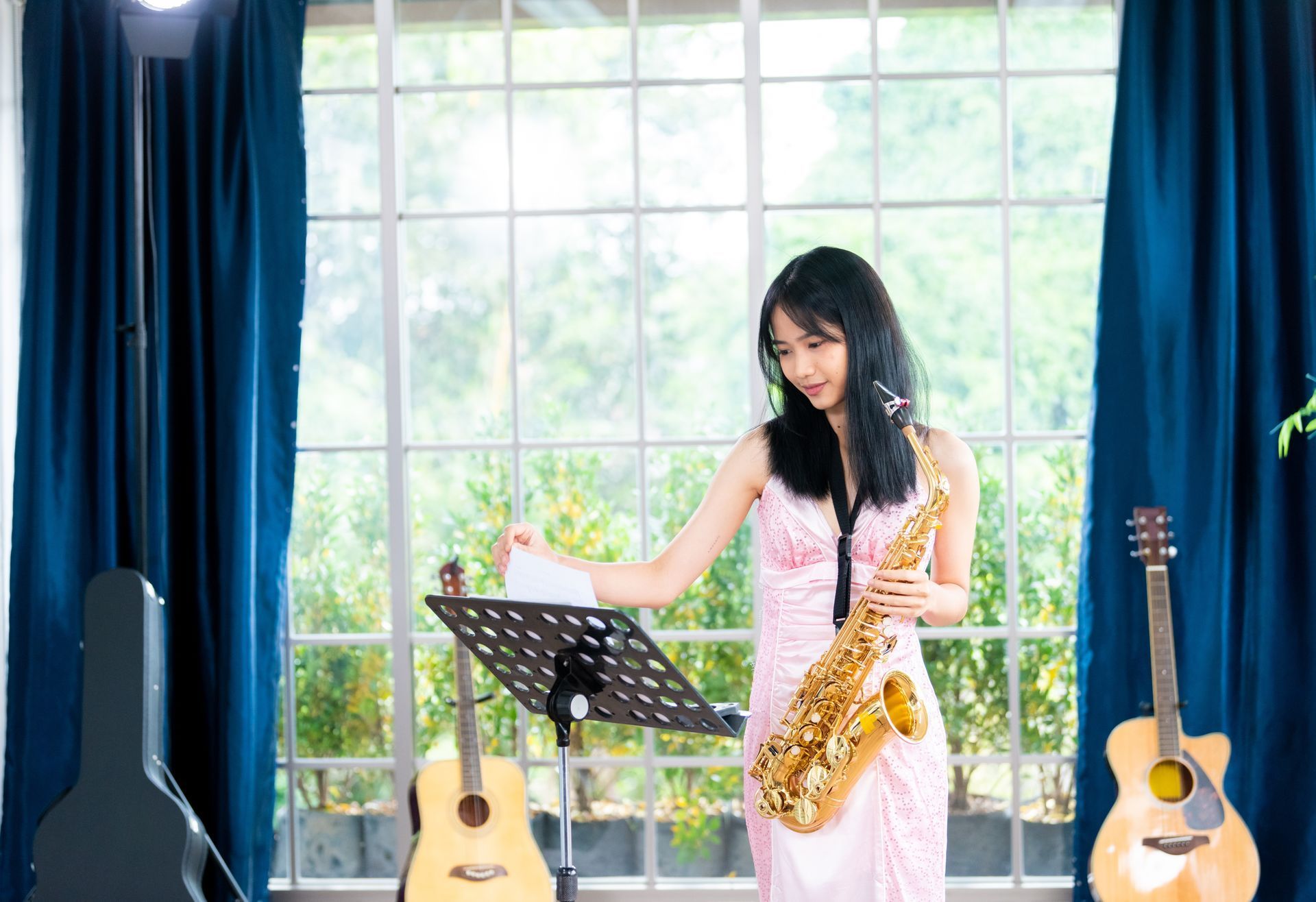 Woman playing saxophone beside music stand in a bright room with guitars and large window.