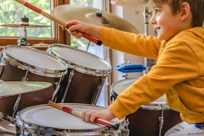 Boy playing drums, smiling, yellow sweatshirt, wooden drumsticks, indoor setting.