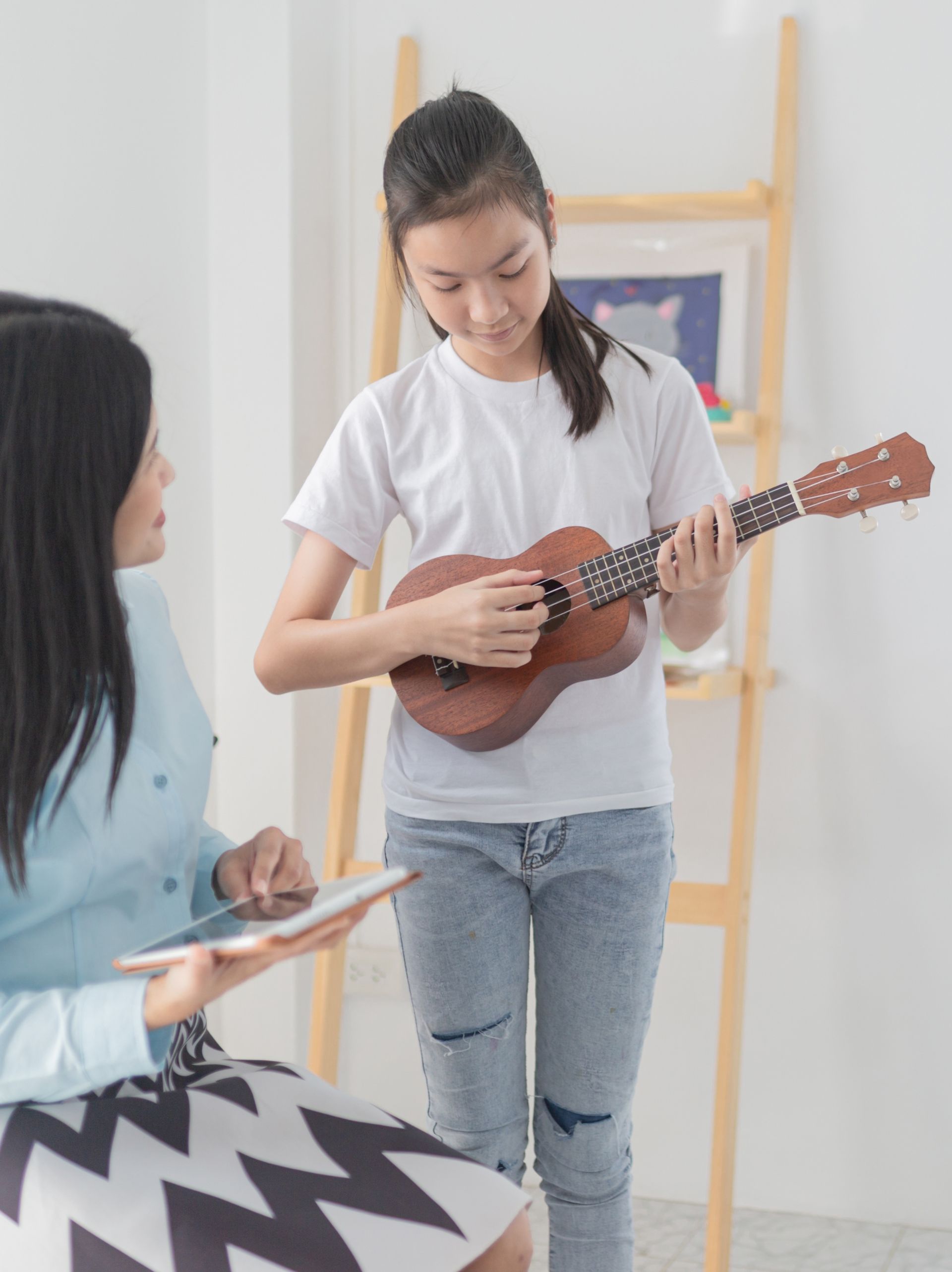 Woman in skirt with tablet watches girl play ukulele in bright room.