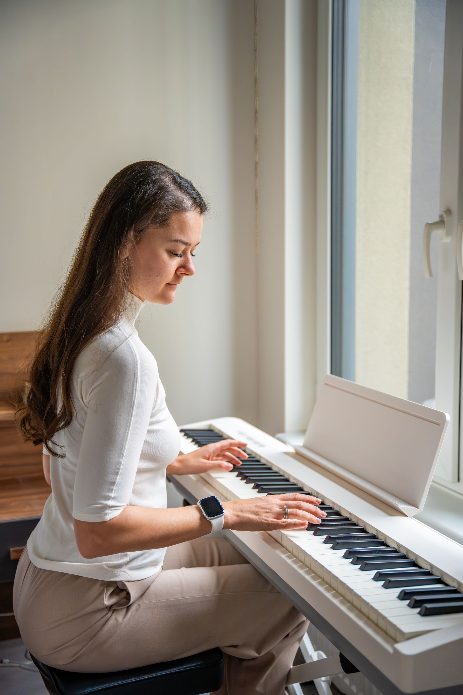 Woman playing a white piano near a window. She's wearing a white top and beige pants.