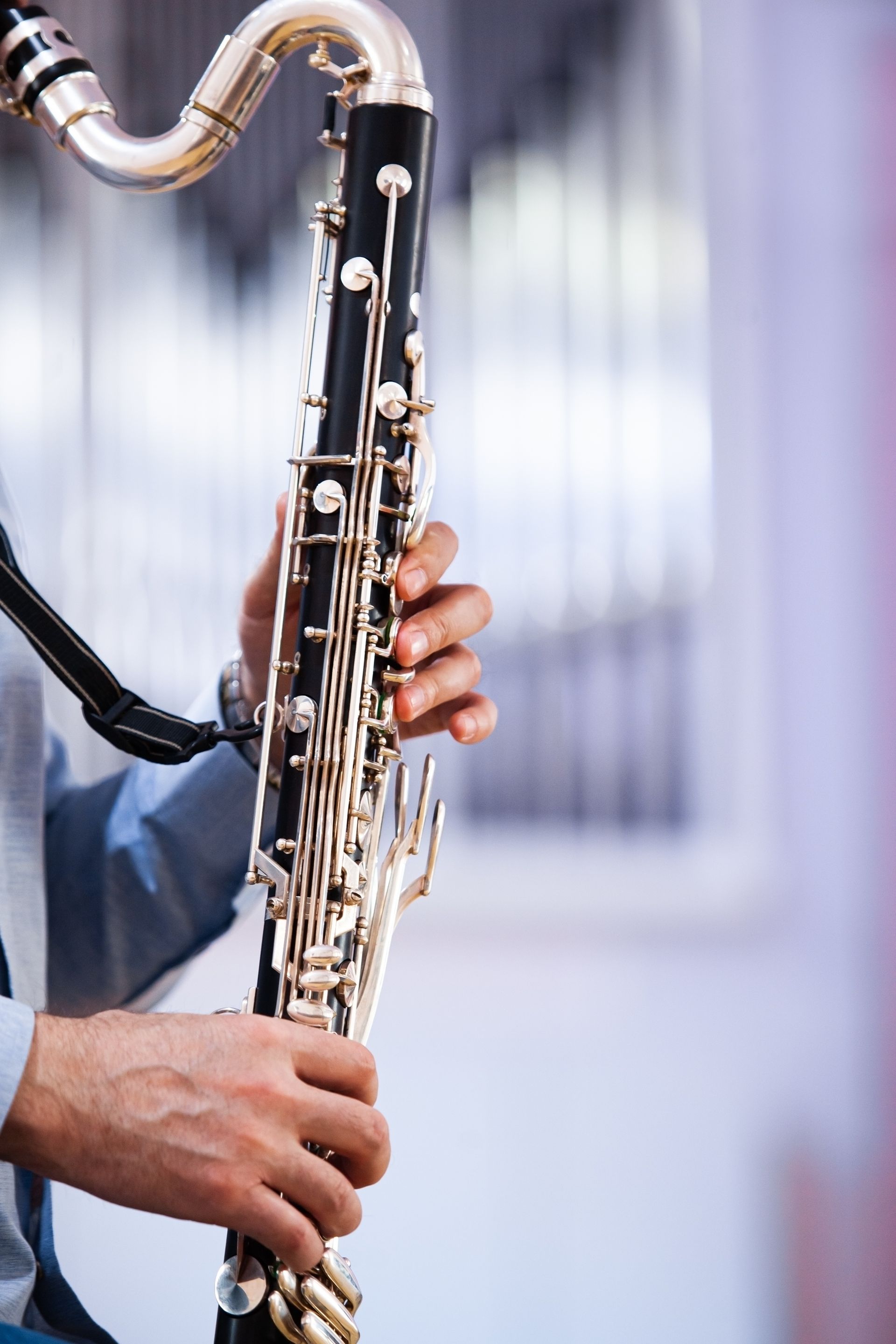 Person playing a bass clarinet. Instrument is black with silver keys, held near a building.