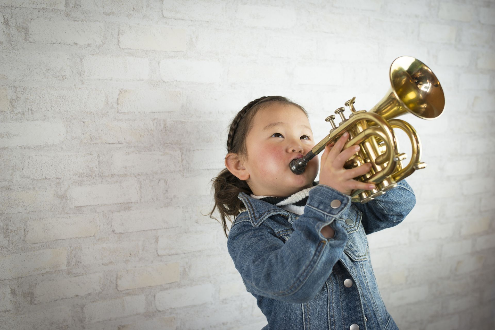 A little girl is playing a trumpet in front of a white brick wall.