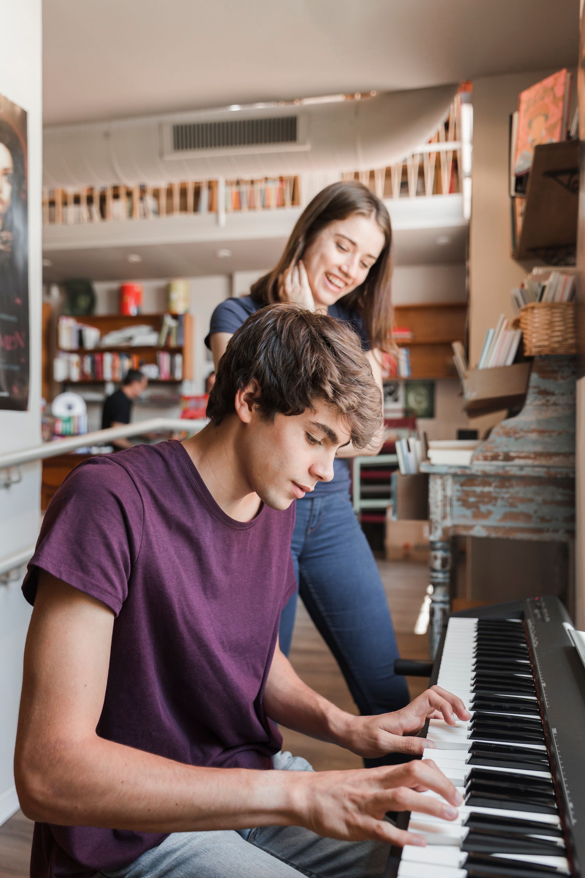 Two people at a piano, one seated and playing while another stands behind in a cozy room.