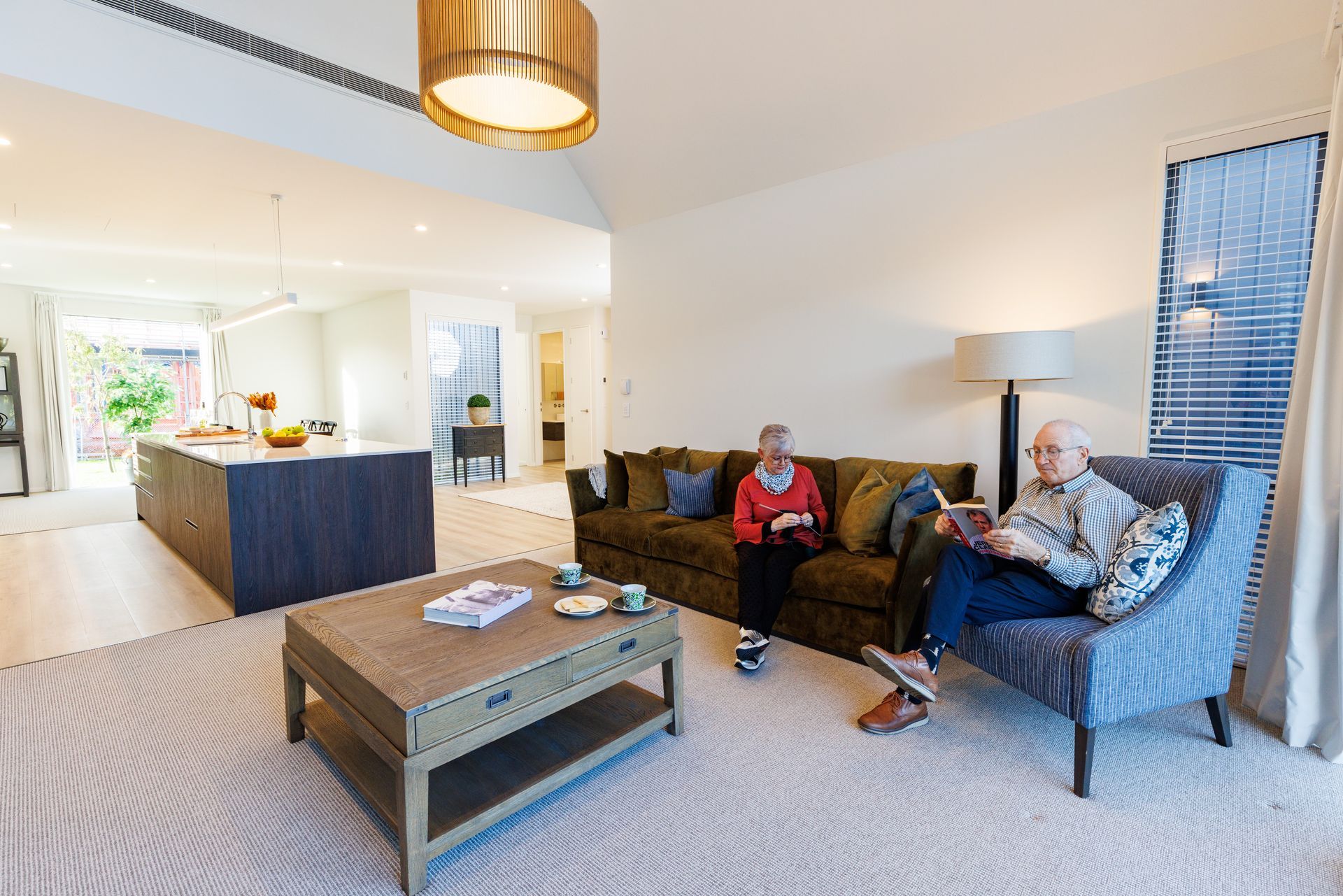 Two people relax in a living room. One sits in an armchair reading a paper, the other sits on a couch using a tablet.