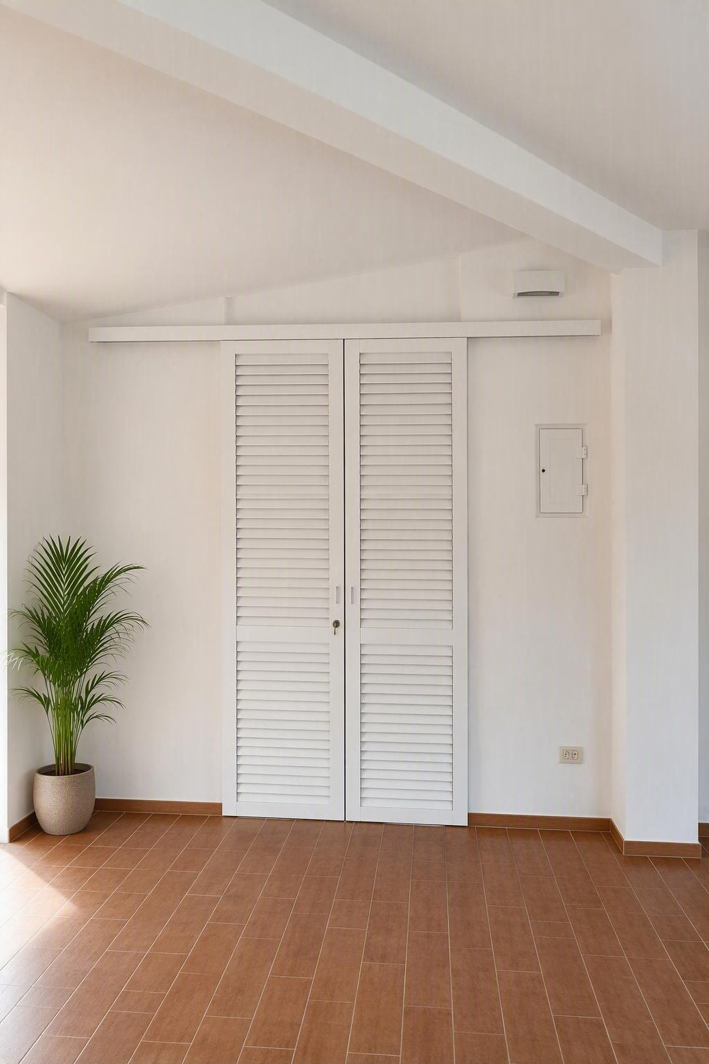 Stanza bianca con soffitto inclinato, doppie porte a persiana chiuse, pavimento in mattoni e una pianta in vaso sulla sinistra.