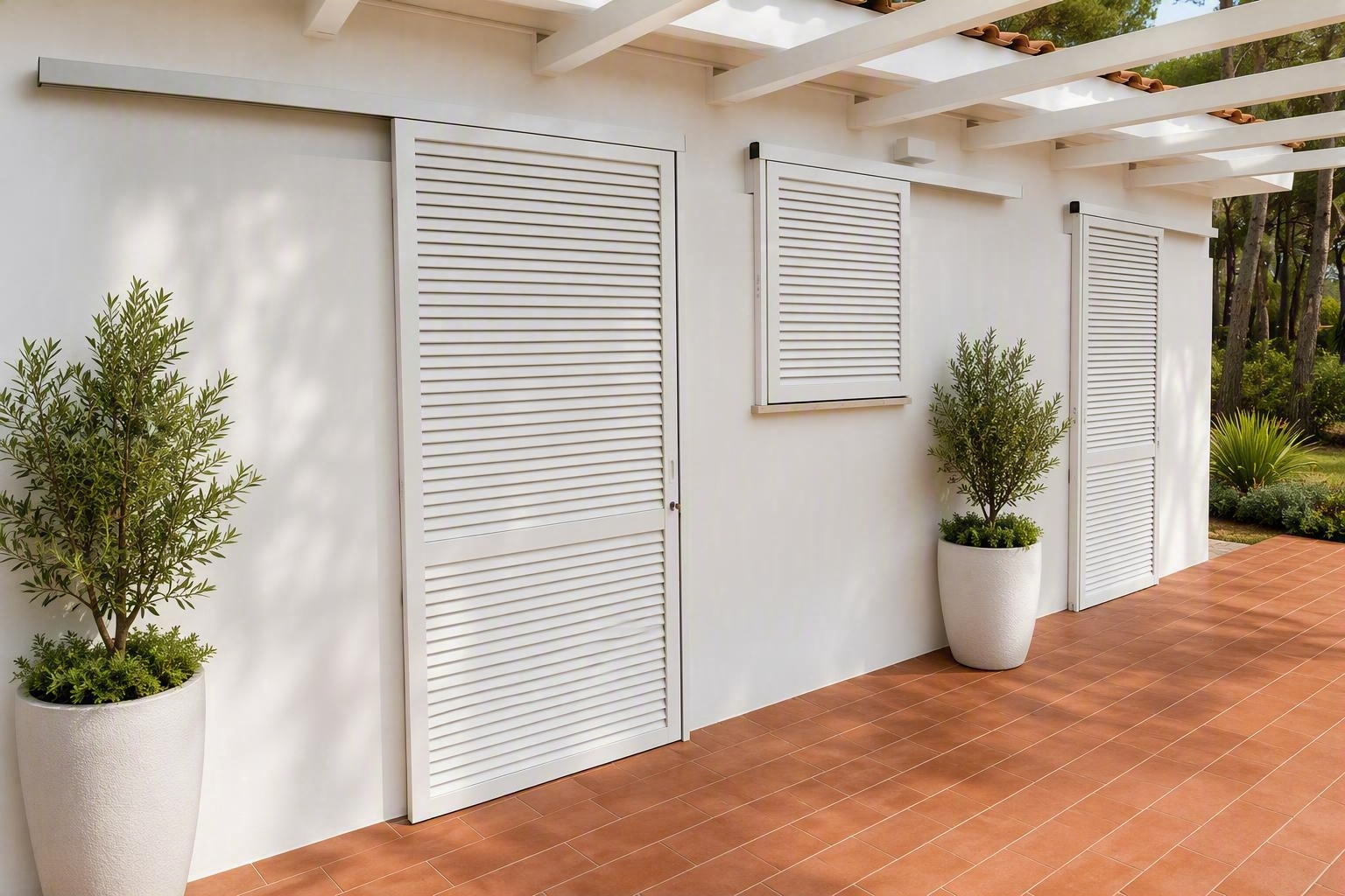 Patio bianco con arbusti in vaso, piastrelle in terracotta e persiane a lamelle sotto un pergolato.