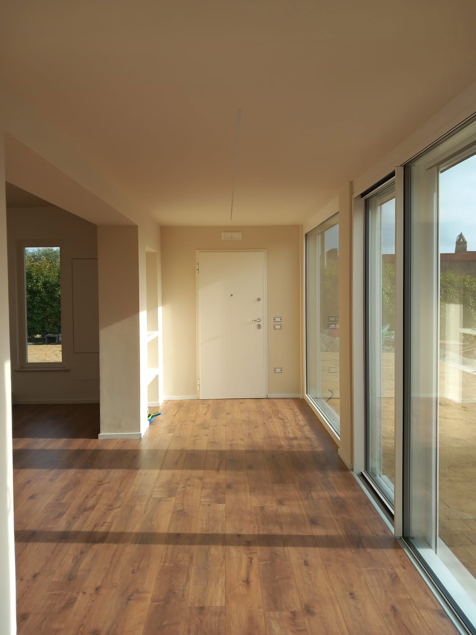 Luminosa stanza vuota con pavimento in legno e grandi porte scorrevoli in vetro lungo la parete destra.