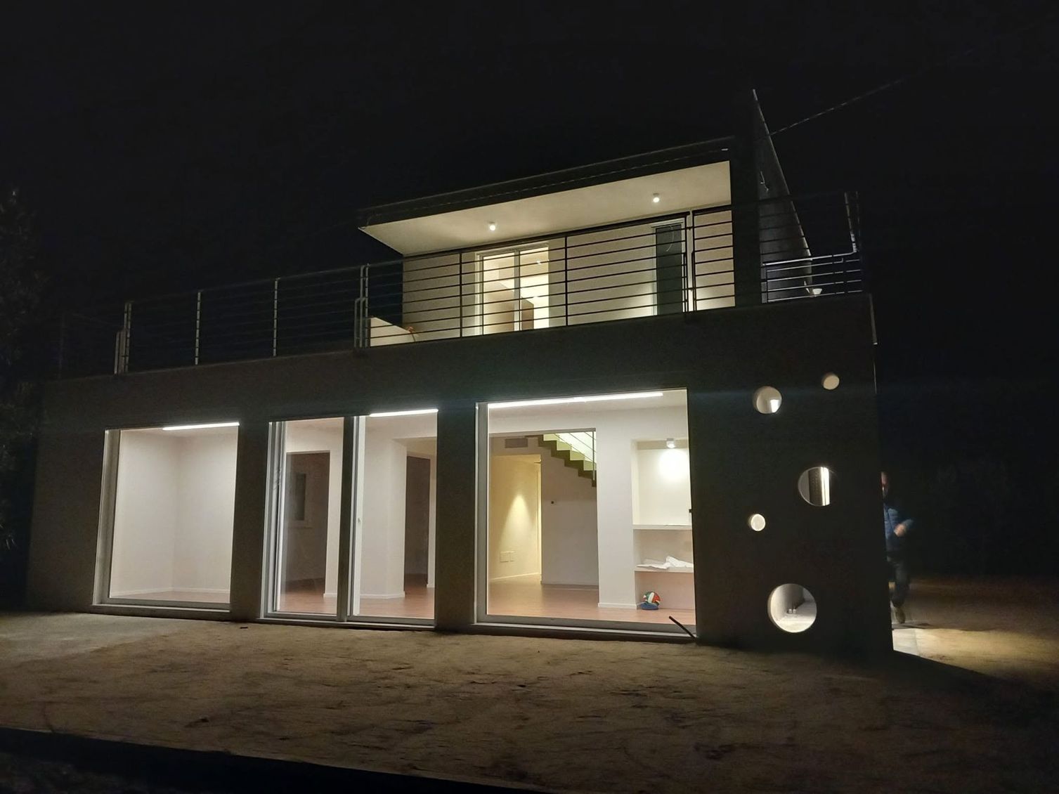 Esterno di una casa moderna di notte, con luci interne brillanti e terrazza sul tetto.