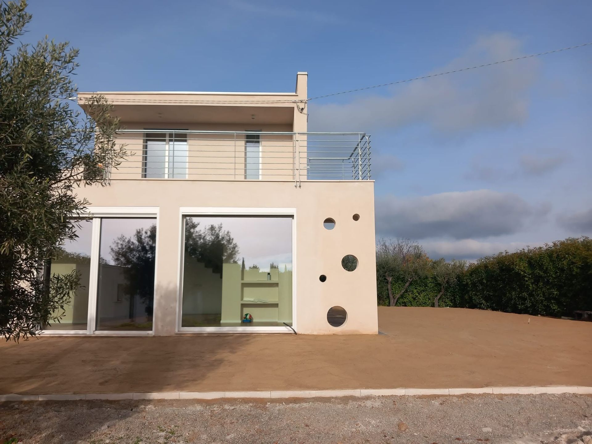 Casa moderna a due piani color beige con grandi porte a vetri, balcone e vialetto fiancheggiato da siepi sotto un cielo nuvoloso.