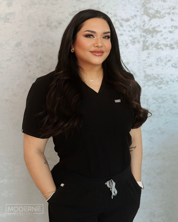 Woman in black scrubs standing against a textured gray wall, hands in pockets, smiling softly.