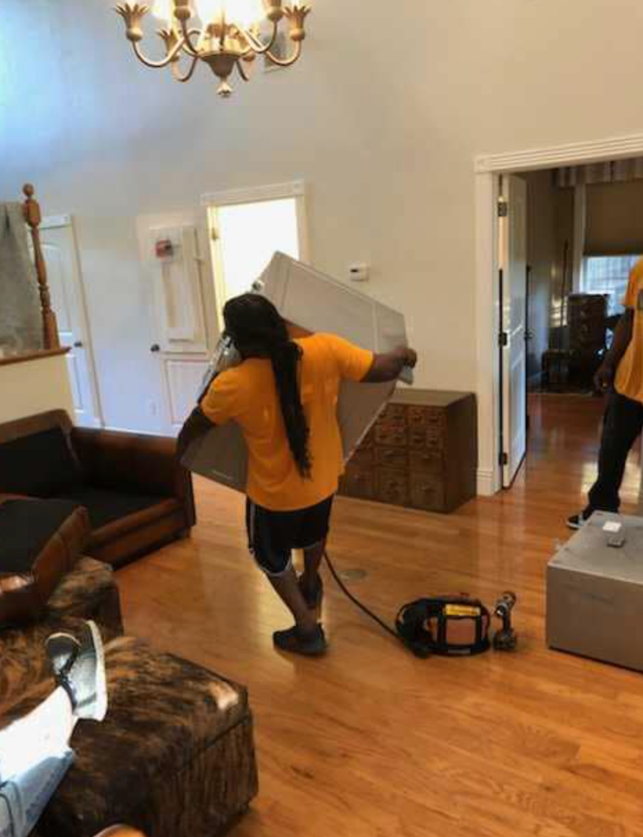 A man in a yellow shirt is carrying a box in a living room.