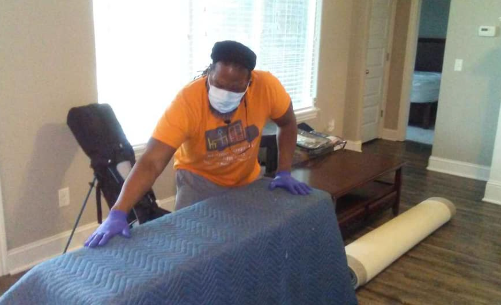 A man wearing a mask and gloves is laying a blue blanket on a table in a living room.