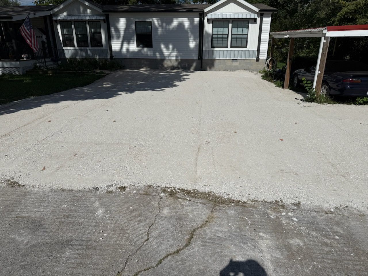 Gravel driveway in front of a white mobile home with a carport on the right and an American flag visible.