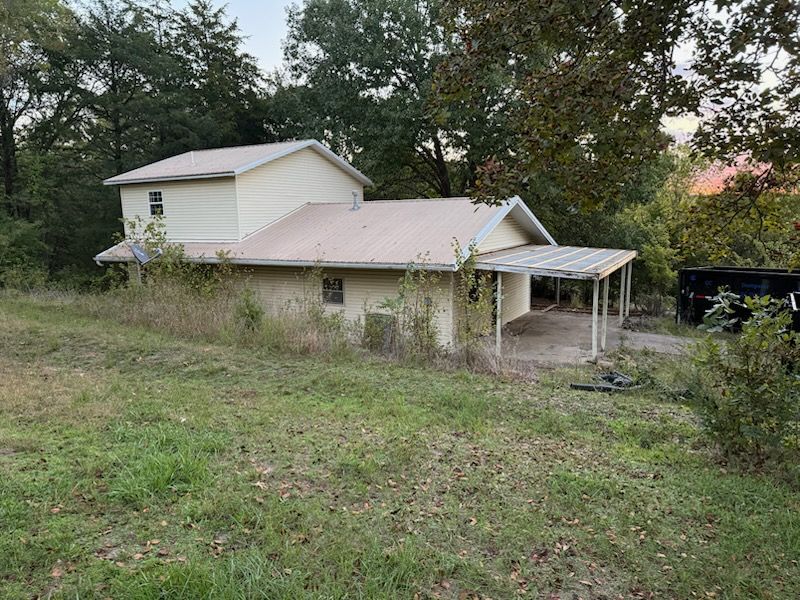 Two-story house with overgrown yard, carport, and metal roof surrounded by trees.