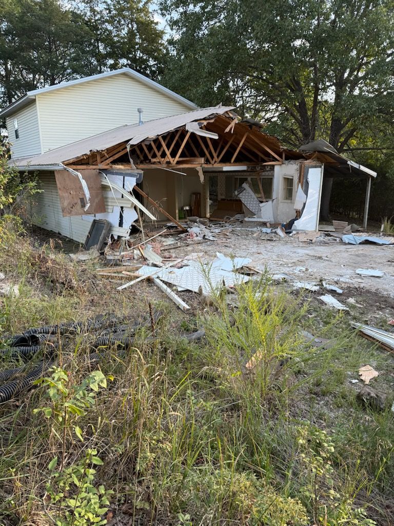 House severely damaged with roof and walls torn away; debris scattered outside.