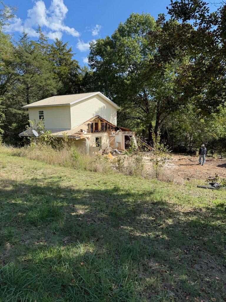 Dilapidated house with missing walls, overgrown yard, man standing nearby.